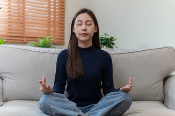 Young Asian woman practicing meditation at home, sitting on a sofa, calm and peaceful. Perfect for...