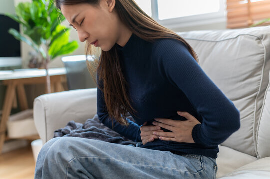 Asian woman sitting on sofa holding her stomach with painful expression, suffering from abdominal pain, stomachache, cramps or digestive issues. Concept for health problems, period pain, gastritis.