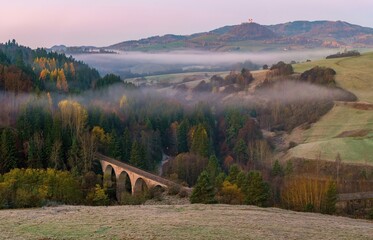 Pink‑tinged dawn light glows over rolling hills and fields of Banská Belá, with low fog drifting through wooded valleys, an old viaduct and autumn colors adding charm to the serene rural scene.