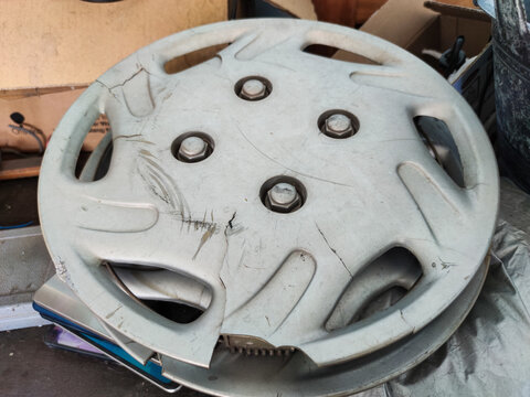 A top-down view of a dirty, gray plastic car hubcap with visible cracks and scratches. The center of the cover features four bolt holes, and the material appears weathered from long-term use.