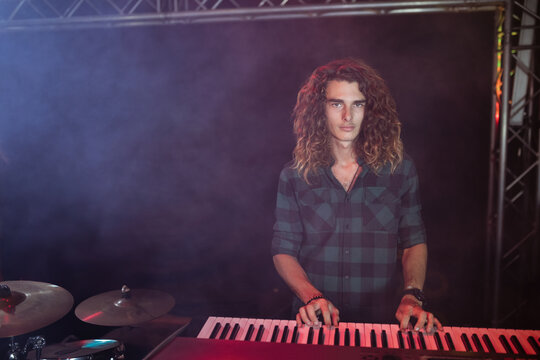 Red keyboard is resting beside drum cymbals on dark stage, illuminated by colored spotlights