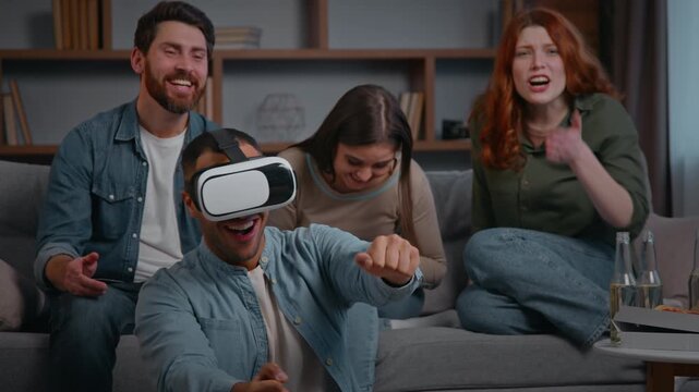 Four multiracial friends at home playing VR game laughing at home party three buddies supporting man in virtual reality helmet play smiling together gaming having fun interactive women men on couch