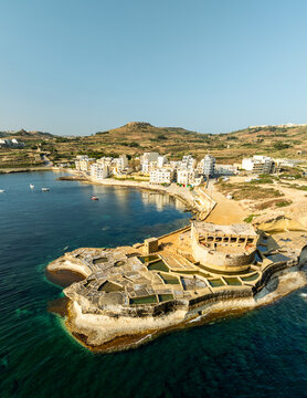 Aerial view of Fort Chambray's weathered stone bastions meet the turquoise sea, contrasting with the distant town under a clear sky, Gozo, Gozo, Malta.