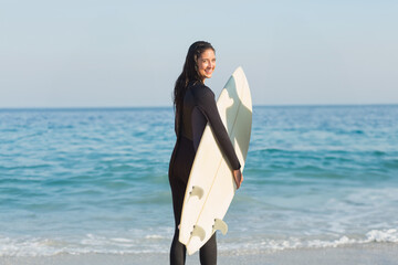 Female surfer wearing black wetsuit smiling while holding white surfboard on sandy beach