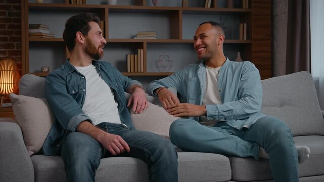 Two multiracial men multiethnic guys talking friendly at home relaxing on couch sofa communicate Caucasian American male friendship discuss funny news smiling carefree conversation in living room