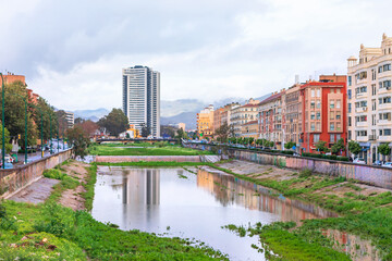 Obraz premium View of Malaga shows a river with reflections, surrounded by colorful historic buildings and a modern tower in distance. Cloudy sky and mountains create a striking urban and natural contrast