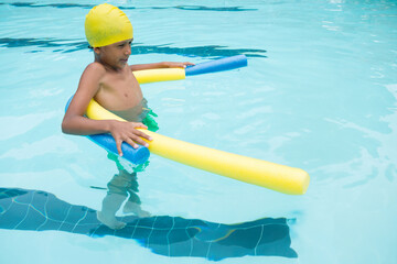 Child boy standing chest-deep in pool wearing yellow swim cap and green trunks holding foam noodles