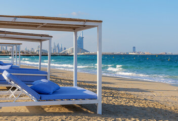 Beach cabanas with blue mattresses on a sandy shore in Dubai. Turquoise sea waves and modern skyline of Dubai in the background. Relaxing tropical vibe of Dubai's coastal lifestyle