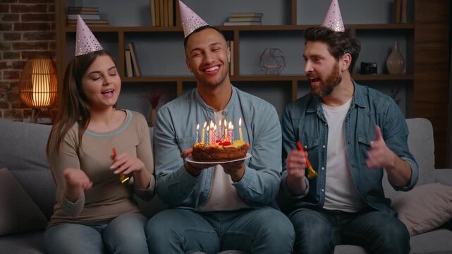Multiracial three friends at home birthday party congratulate American man with cake burning candles smiling laughing happy diversity people surprising African male singing festive song in living room