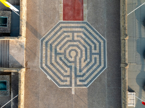 Aerial view of the labyrinth's intricate geometric patterns, a puzzle of stone and shadow, contrasting with the warm, sun-kissed plaza, Gozo, Malta.