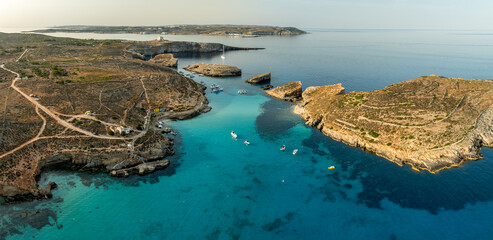 Aerial view of azure waters embracing the rugged, sun-kissed shores of Comino, where boats dance on the surface, revealing the Blue Lagoon's allure, Comino, Malta.
