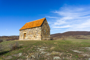 Fototapeta premium Small shurch on the rock. Wooden door. Orange tiled roof, stone walls. Green grass and bushes. Bright blue sly with clouds. Georgia