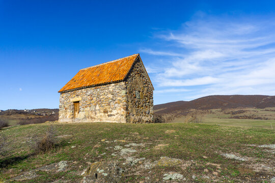 Small shurch on the rock. Wooden door. Orange tiled roof, stone walls. Green grass and bushes. Bright blue sly with clouds. Georgia