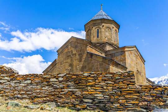 Gergeti Holy Trinity Church. Stone wall fence, dry masonry, orange lichen, stone walls with bas-reliefs, dome with a cross. Bright blue sky with clouds.