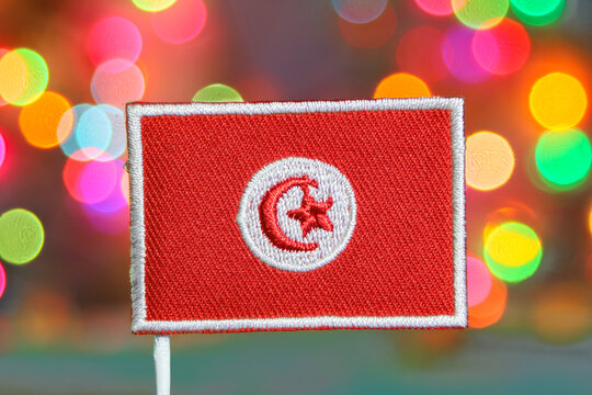 Tunisian embroidered flag in front of colorful bokeh lights, symbolizing New Year celebration, fireworks glow and festive holiday spirit.