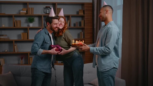 Three multiracial friends at home party birthday congratulation couple Caucasian woman with Latino American male congratulate man giving present box gift cake with candles blowing celebrate together