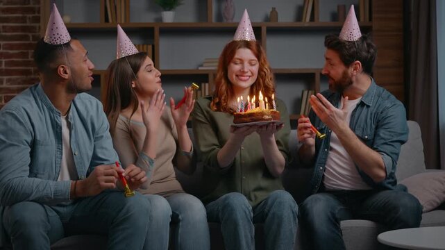 Four multiracial friends singing happy birthday song to Caucasian woman holding festive cake with candles smiling satisfied congratulate girl at home party diverse teammates women men blowing whistles