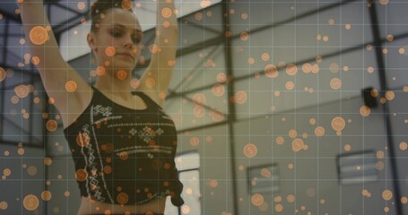 Stretching teenage athlete raising arms overhead in dance studio with beams, windows, orange icons
