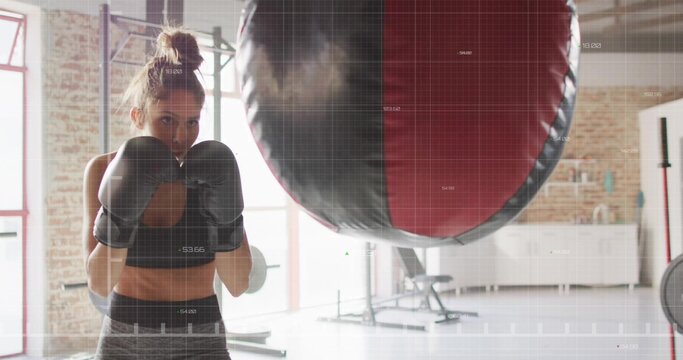 Woman boxer wearing sports bra throwing punches at punching bag in gym studio, with boxing gloves