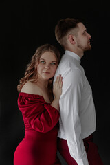 Young Caucasian woman with wavy brown hair in a red dress stands close to a young Caucasian man with short brown hair in a white shirt. They pose against a black background.