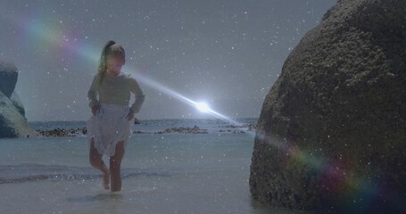Walking child wearing white skirt through shallow seawater at rocky beach, with sunset lens flares