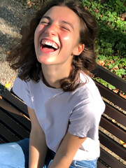 Young woman enjoying a sunny day outdoors, laughing while sitting on a bench in a green park surrounded by trees and nature