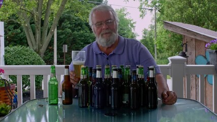 A senior man sits at a table on a patio, surrounded by an assortment of beer bottles. He relaxes while sipping from a glass and enjoys a peaceful afternoon in a lush, green setting.