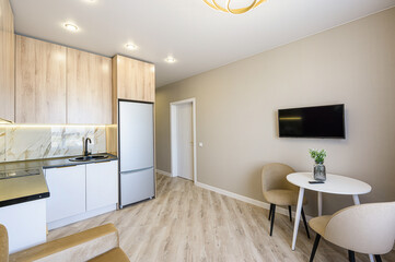 modern apartment interior with a kitchen area featuring wooden cabinets and a dining area with a white table and chairs. A TV is mounted on the wall