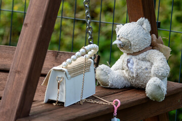 Teddy bear with a purse sitting on a wooden swing in a garden setting
