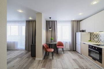 bright, modern apartment interior featuring a kitchen area with light wood floors, beige cabinets, and pink chairs around a small table. Large windows let in natural light