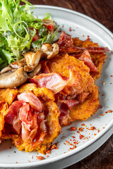 Rustic meal on white plate: crispy potato pancakes with bacon, sautéed mushrooms, fresh salad, creamy dip, and crushed chili flakes. Top view on dark wooden background