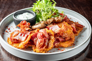 Rustic meal on white plate: crispy potato pancakes with bacon, sautéed mushrooms, fresh salad, creamy dip, and crushed chili flakes. Top view on dark wooden background