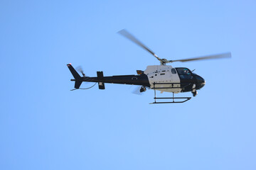 Fototapeta premium Black and white helicopter flying against a clear blue sky, captured in motion with spinning rotor blades.