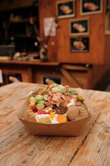Craft box with oven-baked potato filled with tuna, olives, lime, fresh herbs, and creamy sauce, placed on a wooden outdoor table, with a wooden stall featuring artwork on shutters in the background.