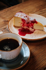 Cup of black coffee on saucer served with cheesecake drizzled with red berry sauce on a wooden table, creating cozy and inviting café dessert scene.