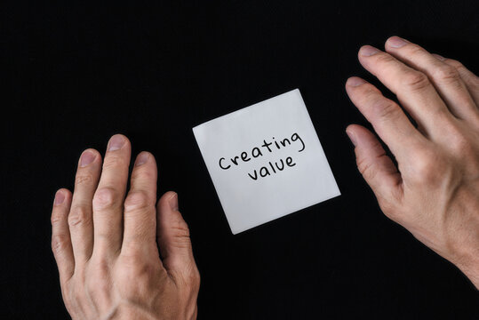 Naklejki Male hands and memo paper with text "Creating value" isolated on black background. Business strategy essentials value creation note on black, minimal flat lay concept