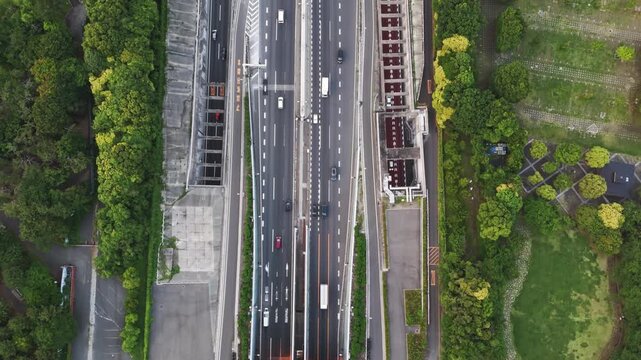 Aerial view of a multilane expressway entering a tunnel by a green city park in Odaiba. Cars stream in both directions, lane marking and barriers forming crisp geometry that reads like a living map