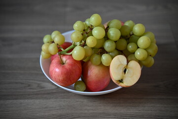 A fresh assortment of green grapes and red apples, one apple sliced ​​to show its core, in a white bowl on a wooden table. The composition is bright and natural.