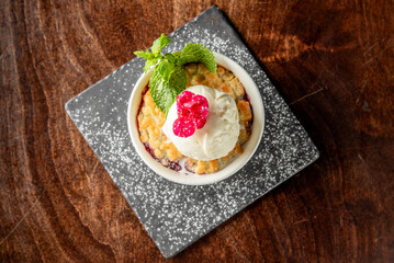 Fruit crumble dessert with vanilla ice cream, mint, and berries in a white ramekin on a black plate dusted with powdered sugar. Rustic wooden background, top view