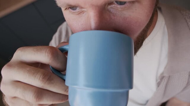 Early morning from above scene of Caucasian bearded man taking sip of hot, freshly brewed coffee from blue mug in kitchen