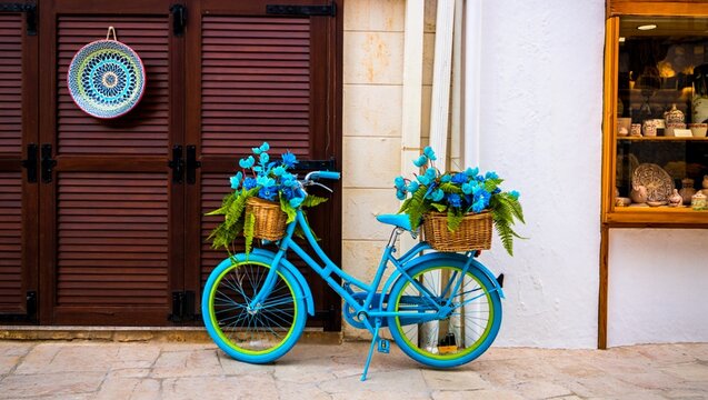 A charming turquoise vintage bicycle adorned with vibrant blue flowers and green ferns in wicker baskets, parked against a quaint European building facade with wooden shutters and a shop window displa
