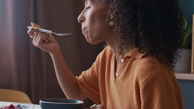 Side-view closeup of young Biracial woman enjoying cornflakes with yogurt and fresh berries in bowl, capturing healthy breakfast moment at home