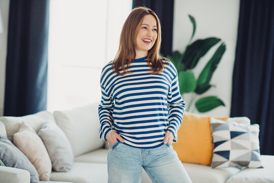 Charming woman in a striped sweatshirt standing confidently in a cozy living room with modern decor and soft lighting.