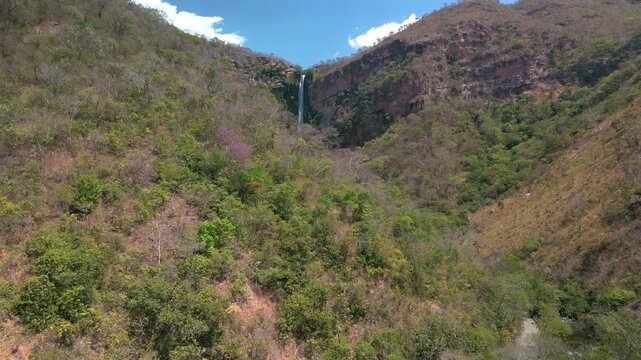 A imagem a&eacute;rea revela uma imponente cachoeira que despenca de um pared&atilde;o rochoso envolto por vegeta&ccedil;&atilde;o t&iacute;pica do cerrado, mesclando tons terrosos das pedras e verdes vibrantes das copas das &aacute;rvores.