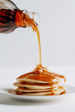 ottle pouring maple syrup directly onto pancakes on a white plate. The bottle is held at a certain distance above the pancakes so that the maple syrup drips in a long stream. High-quality photo, white