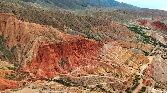 Panoramic drone shot of Skazka Canyon in Kyrgyzstan, showing a surreal red and yellow landscape with rocky formations under dramatic skies.