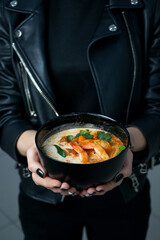 Tom yam in a black deep plate in the hands of a girl in a jacket