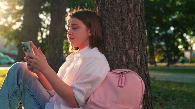 Schoolgirl girl sits with smartphone under tree on lawn in park. Education. Teenager schoolboy with school backpack uses smartphone while sitting in city park. Child relaxes with gadget natural park - Powered by Adobe