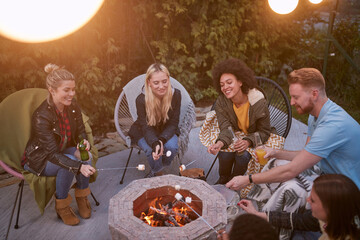 Young adults sitting by the fire on an autumn afternoon, roasting marshmallows and drinking beer together.