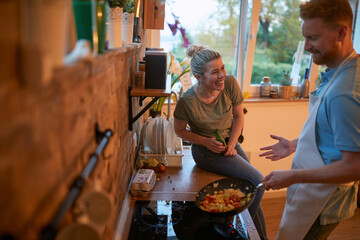 Man enjoying time with girlfriend and making dinner in the kitchen, roasting vegetables in a pan.